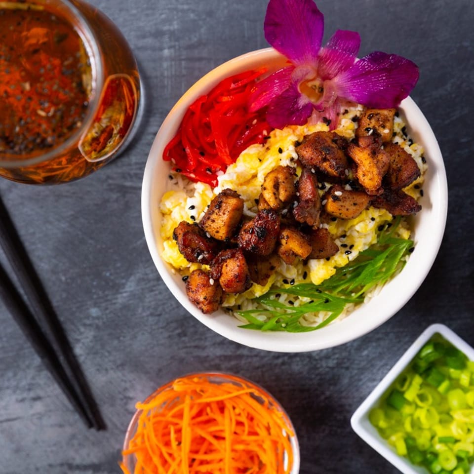Bowl of rice topped with grilled meat, vegetables, and edible flower, surrounded by chopsticks, sauces, corn, carrots, and scallions.