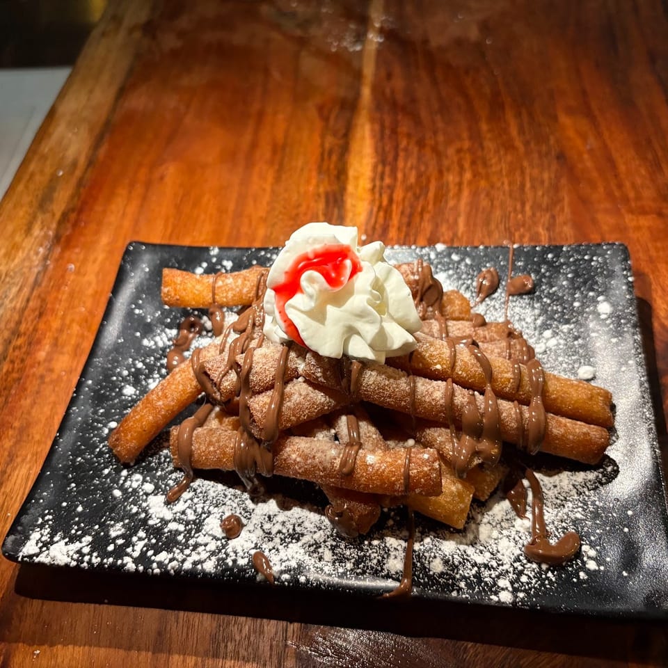 Crispy churros piled on a black plate, drizzled with chocolate, dusted with sugar, and topped with whipped cream and strawberry sauce.